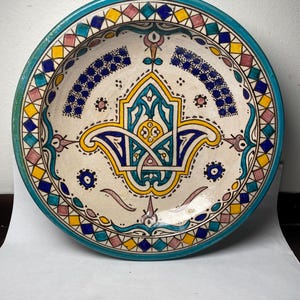 May include: A decorative ceramic plate with a white base and intricate hand-painted designs. The plate features a turquoise rim and a central motif in yellow, blue, and teal. The border includes geometric patterns in yellow, pink, and blue. A unique piece for home decor.