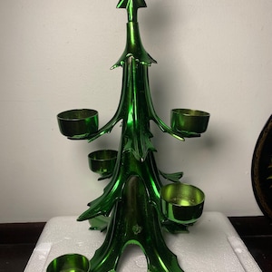 May include: A metallic green Christmas tree-shaped candle holder featuring a star topper and five circular candle cups extending from the branches. The structure is made of a smooth, reflective material with a vibrant, glossy finish throughout.
