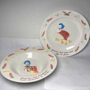 May include: Two white ceramic plates with a duck and chick design. The plates have a border of carrots, eggs, and flowers. One plate reads "Gemima laid some more eggs in June" and the other reads "Gemima was burdened with bunches of herbs."