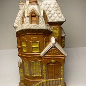 May include: A ceramic house figurine with a brown body, white roof, and yellow window accents. The house features a detailed facade with a front porch and chimney. The overall design is reminiscent of a Victorian-style home.