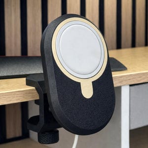 K&ouml;nnte beinhalten: Eine schwarz-goldene magnetische Ladestation mit einer kreisf&ouml;rmigen wei&szlig;en Mitte, die eine strukturierte Oberfl&auml;che und einen stabilen Klemmmechanismus mit einem ger&auml;ndelten Einstellknopf aufweist, um sicher an einer Tischkante zu befestigen.