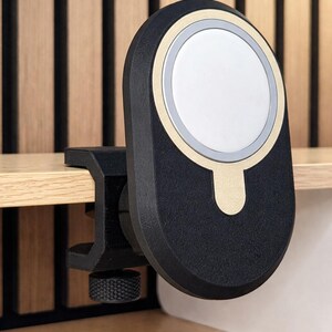 K&ouml;nnte beinhalten: Eine magnetische Ladestation in Schwarz und Gold mit einem kreisf&ouml;rmigen wei&szlig;en Mittelpad. Das Ger&auml;t verf&uuml;gt &uuml;ber einen schwarzen C-Klemmen-Mechanismus mit einem strukturierten Einstellknopf, der zur Befestigung an einer flachen Kante dient.