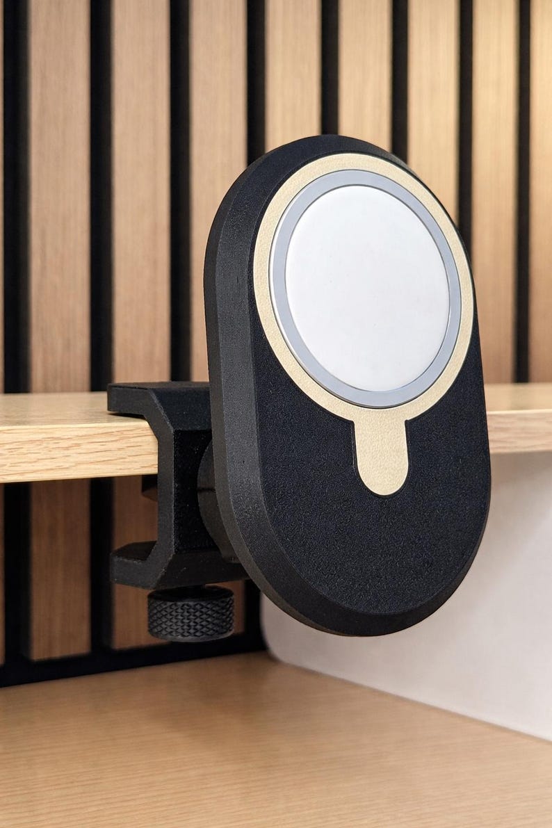 K&ouml;nnte beinhalten: Eine magnetische Ladestation in Schwarz und Gold mit einem kreisf&ouml;rmigen wei&szlig;en Mittelpad. Das Ger&auml;t verf&uuml;gt &uuml;ber einen schwarzen C-Klemmen-Mechanismus mit einem strukturierten Einstellknopf, der zur Befestigung an einer flachen Kante dient.