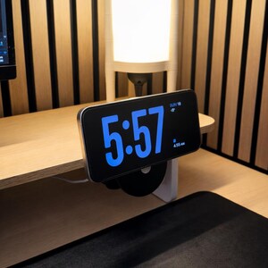 K&ouml;nnte beinhalten: Ein Smartphone auf einem schwarzen, runden St&auml;nder zeigt eine digitale Uhr mit gro&szlig;en blauen Ziffern, die 5:57 Uhr anzeigen, dazu das Datum SUN 18 und ein kleines Weckersymbol mit dem Text 4:55 AM. Das Ger&auml;t ist horizontal aufgestellt.