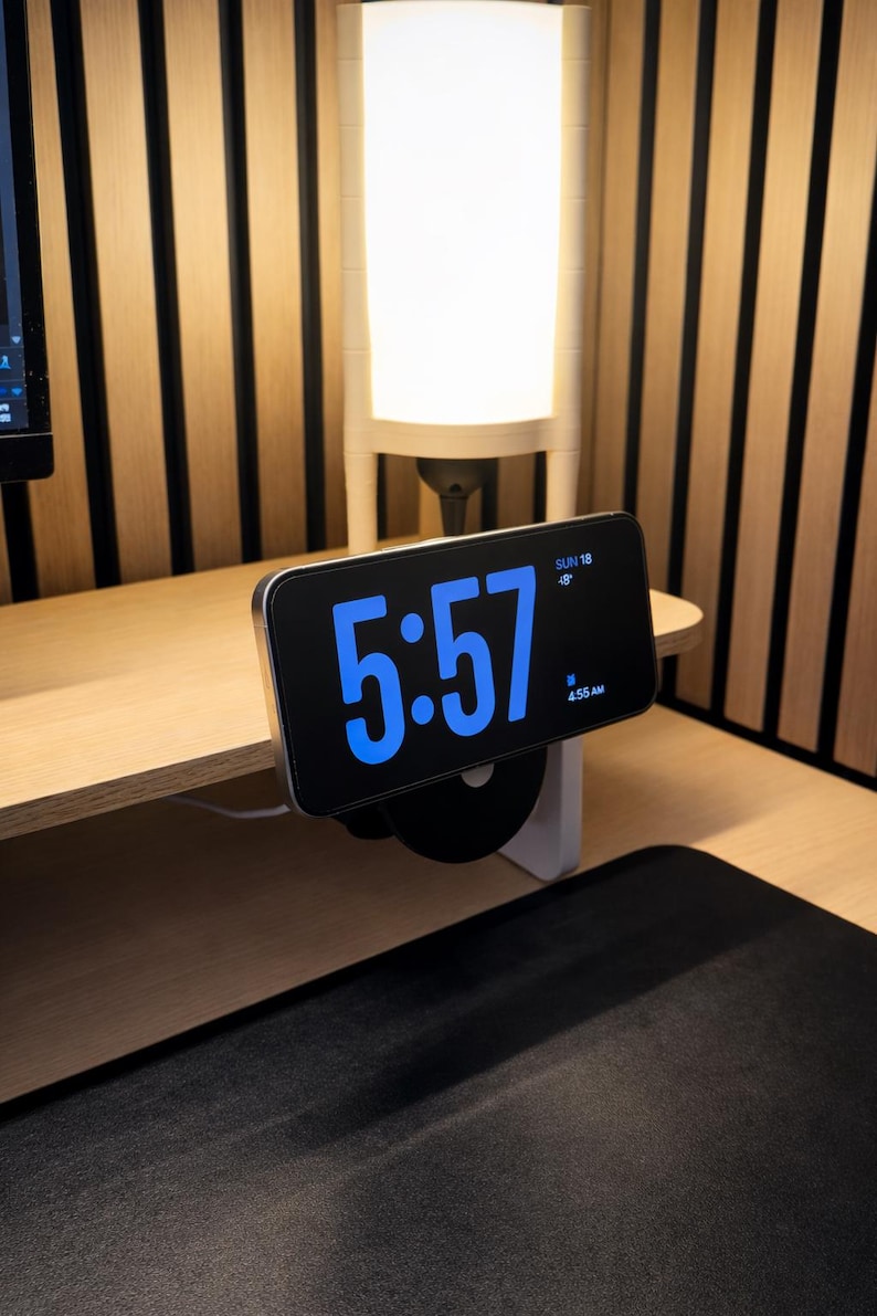 K&ouml;nnte beinhalten: Ein Smartphone auf einem schwarzen, runden St&auml;nder zeigt eine digitale Uhr mit gro&szlig;en blauen Ziffern, die 5:57 Uhr anzeigen, dazu das Datum SUN 18 und ein kleines Weckersymbol mit dem Text 4:55 AM. Das Ger&auml;t ist horizontal aufgestellt.