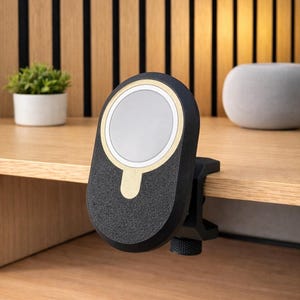 May include: A black, oval-shaped magnetic charging mount with a gold-colored circular accent and a central white disc. The device features a textured finish and is attached to the edge of a wooden surface using a black clamp with a tightening knob.