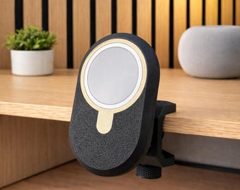 Op de afbeelding: Een zwarte, ovaalvormige magnetische oplaadhouder met een goudkleurig cirkelvormig accent en een witte schijf in het midden. Het apparaat heeft een getextureerde afwerking en is bevestigd aan de rand van een houten oppervlak met een zwarte klem.