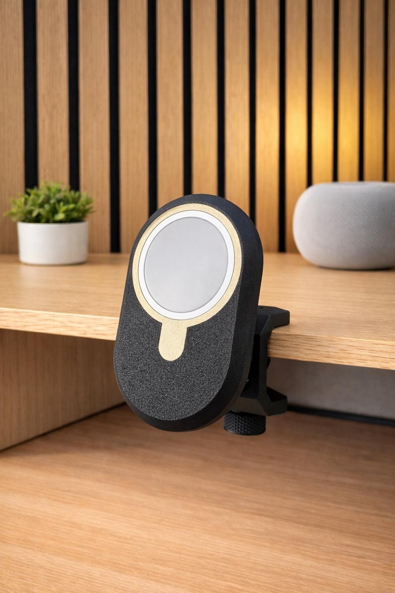 K&ouml;nnte beinhalten: Eine schwarze, oval geformte magnetische Ladestation mit einem goldfarbenen kreisf&ouml;rmigen Akzent und einer wei&szlig;en Scheibe in der Mitte. Das Ger&auml;t hat eine strukturierte Oberfl&auml;che und ist mit einer schwarzen Klemme mit Drehknopf an einer Holzkante befestigt.