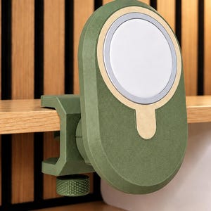 K&ouml;nnte beinhalten: Eine magnetische Handyhalterung in mattem Olivgr&uuml;n mit einer kreisf&ouml;rmigen wei&szlig;en Ladefl&auml;che und einem goldfarbenen Akzentring. Das Ger&auml;t verf&uuml;gt &uuml;ber einen stabilen Klemmmechanismus mit einem strukturierten gr&uuml;nen Drehknopf zur Befestigung.