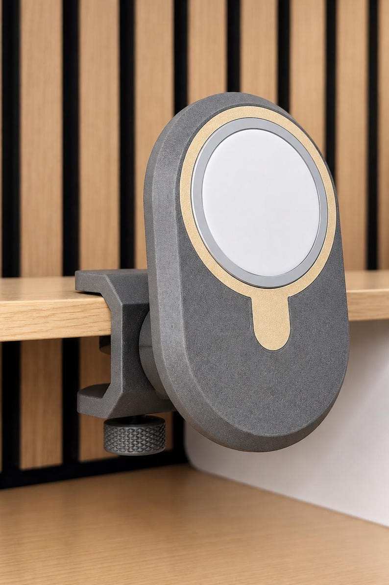 K&ouml;nnte beinhalten: Eine dunkelgraue, ovale magnetische Ladestation mit einem goldfarbenen kreisf&ouml;rmigen Akzent und einer wei&szlig;en Mitte. Das Ger&auml;t verf&uuml;gt &uuml;ber einen stabilen Klemmmechanismus mit einem strukturierten Einstellknopf zur Befestigung an einer Kante.