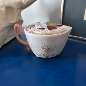 May include: A set of four white ceramic measuring cups with a pink rim and a Tinkerbell illustration. The cups are nested inside each other.