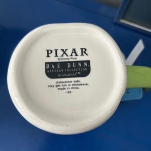 May include: White ceramic mug bottom with the text "PIXAR" and "RAE DUNN. ARTISAN COLLECTION BY MAGENTA". The text "dishwasher safe. may get hot in microwave. made in china." is also visible. The mug is part of the Disney/Pixar collection.