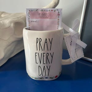 Puede incluir: Taza de cerámica blanca con las palabras "PRAY EVERY DAY" en letras negras. La taza contiene una pila de tarjetas "PRAYERS" rosas y blancas. Una cinta blanca y una etiqueta están unidas al asa de la taza. Acentos florales decoran la base de la taza.