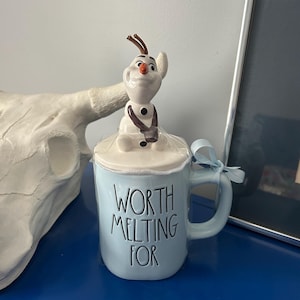 May include: A light blue ceramic mug with the text "WORTH MELTING FOR" in black. The mug features a lid with a figurine of Olaf from Frozen. A light blue ribbon is tied to the handle.