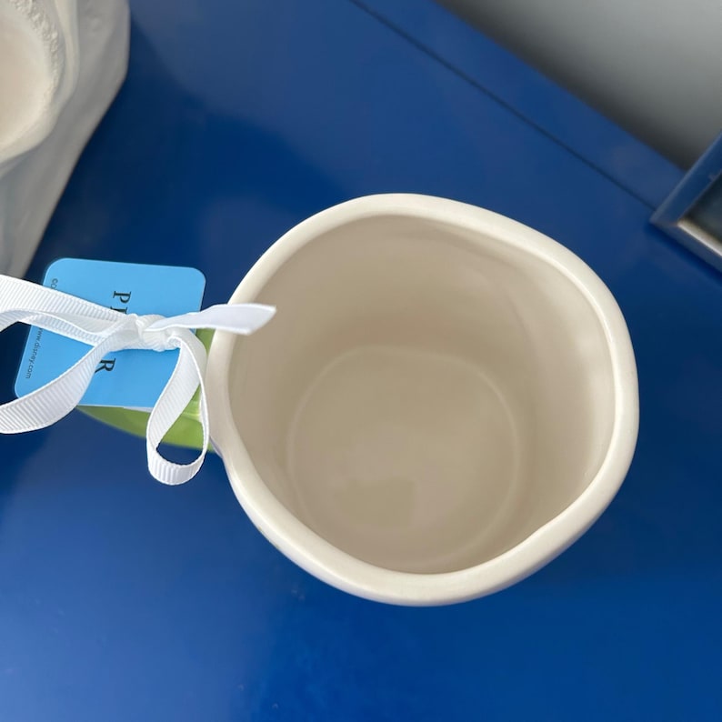 May include: Cream-colored ceramic mug with a rounded shape and a wide opening. A white ribbon is tied around a tag attached to the mug. The mug is set against a blue surface.