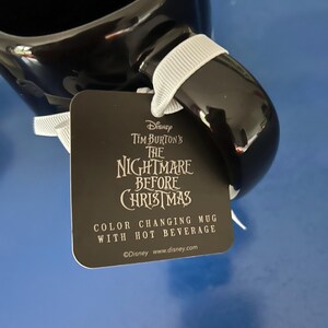 Op de afbeelding: Een zwarte keramische mok met een wit lint aan het handvat. Aan het lint hangt een vierkant label met de tekst: Disney Tim Burton's The Nightmare Before Christmas, Color Changing Mug With Hot Beverage, en Disney www.disney.com.