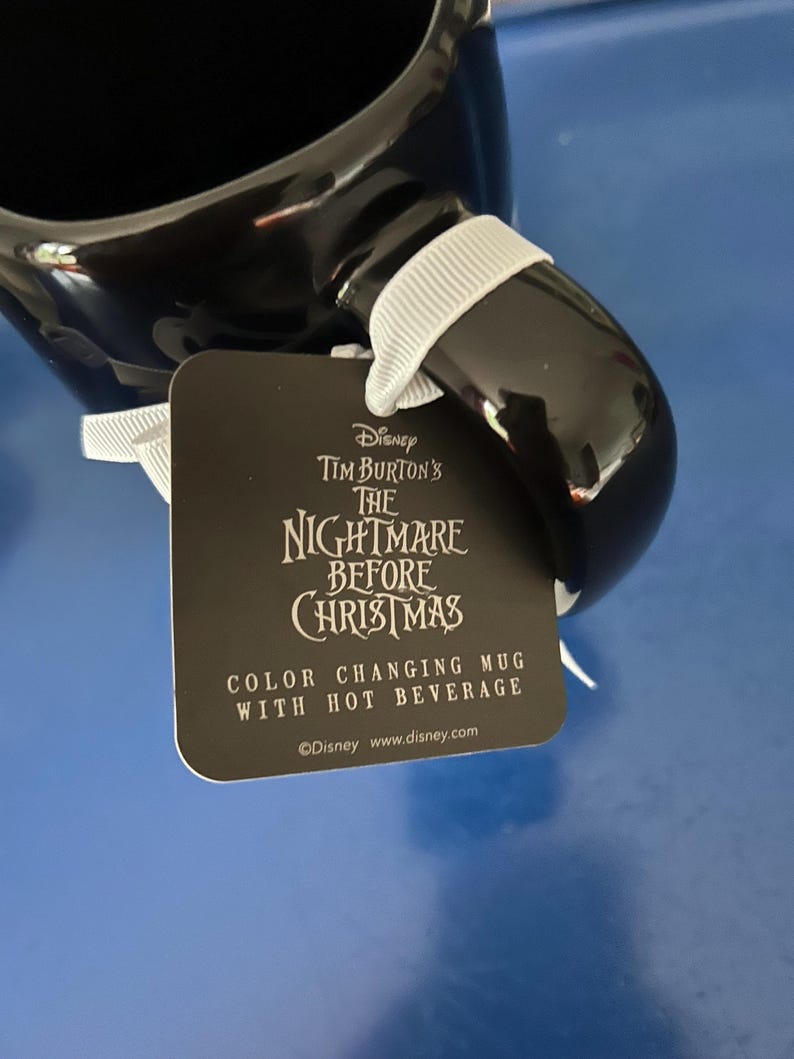Op de afbeelding: Een zwarte keramische mok met een wit lint aan het handvat. Aan het lint hangt een vierkant label met de tekst: Disney Tim Burton's The Nightmare Before Christmas, Color Changing Mug With Hot Beverage, en Disney www.disney.com.