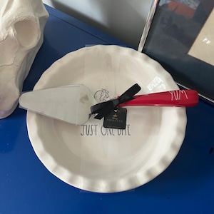 May include: A white ceramic pie dish with a scalloped edge and the black text "JUST ONE BITE" printed inside. A metal pie server with a red handle featuring the white text "YUM" is tied to the dish with a black ribbon and a Disney Princess tag.