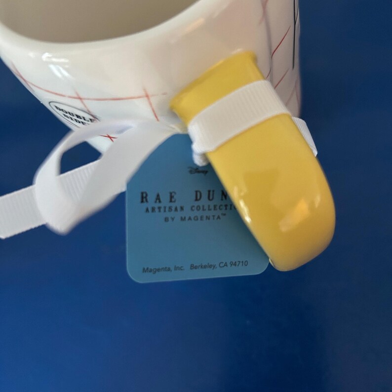 May include: A yellow ceramic mug with a white and red checkered pattern. The mug has a yellow handle with a white stripe. The mug is labeled "Rae Dunn Artisan Collection by Magenta" and "Magenta, Inc. Berkeley, CA 94710".