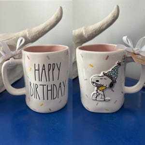 May include: White ceramic mug with a pink interior, decorated with colorful confetti and the words "HAPPY BIRTHDAY" in black. The other side features a Snoopy graphic wearing a party hat. A white ribbon is tied around the handle.