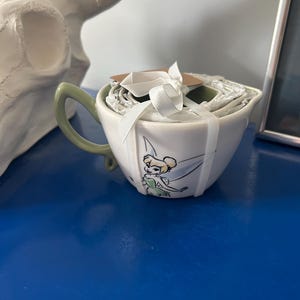 Könnte beinhalten: Ein weißes Messbecher-Set aus Keramik mit einer Tinkerbell-Grafik auf der Vorderseite. Die Becher sind ineinander gestapelt, mit einem weißen Band zusammengebunden und haben einen hellgrünen Griff. Das Set verfügt über einen Ausgießer.
