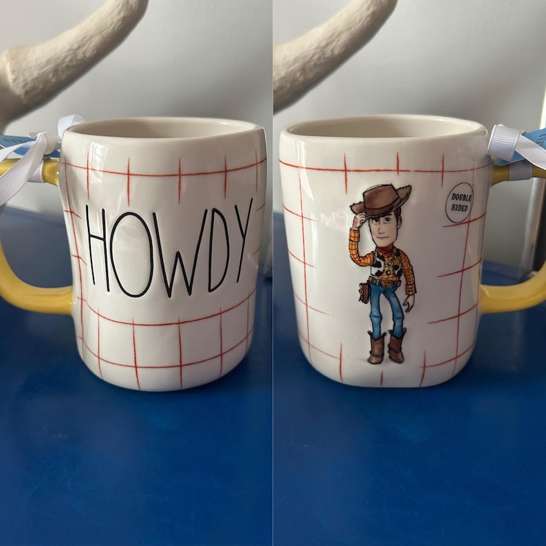 May include: A white ceramic mug with a red and white checkered pattern. The mug has a yellow handle and says "HOWDY" on one side. The other side features a cartoon image of Woody from Toy Story wearing a cowboy hat and holding a lasso. The mug says "DOUBLE SIDED" on the side.