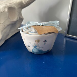 May include: A white ceramic bowl with a light blue rim and a Cinderella graphic. The bowl is tied with a light blue ribbon and has a brown tag attached. The bowl is sitting on a blue surface.