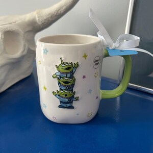 May include: White ceramic mug with a green handle featuring three green alien characters stacked on top of each other. The mug is decorated with colorful star shapes and the text "DOUBLE SIZED". A white ribbon is tied around the handle.