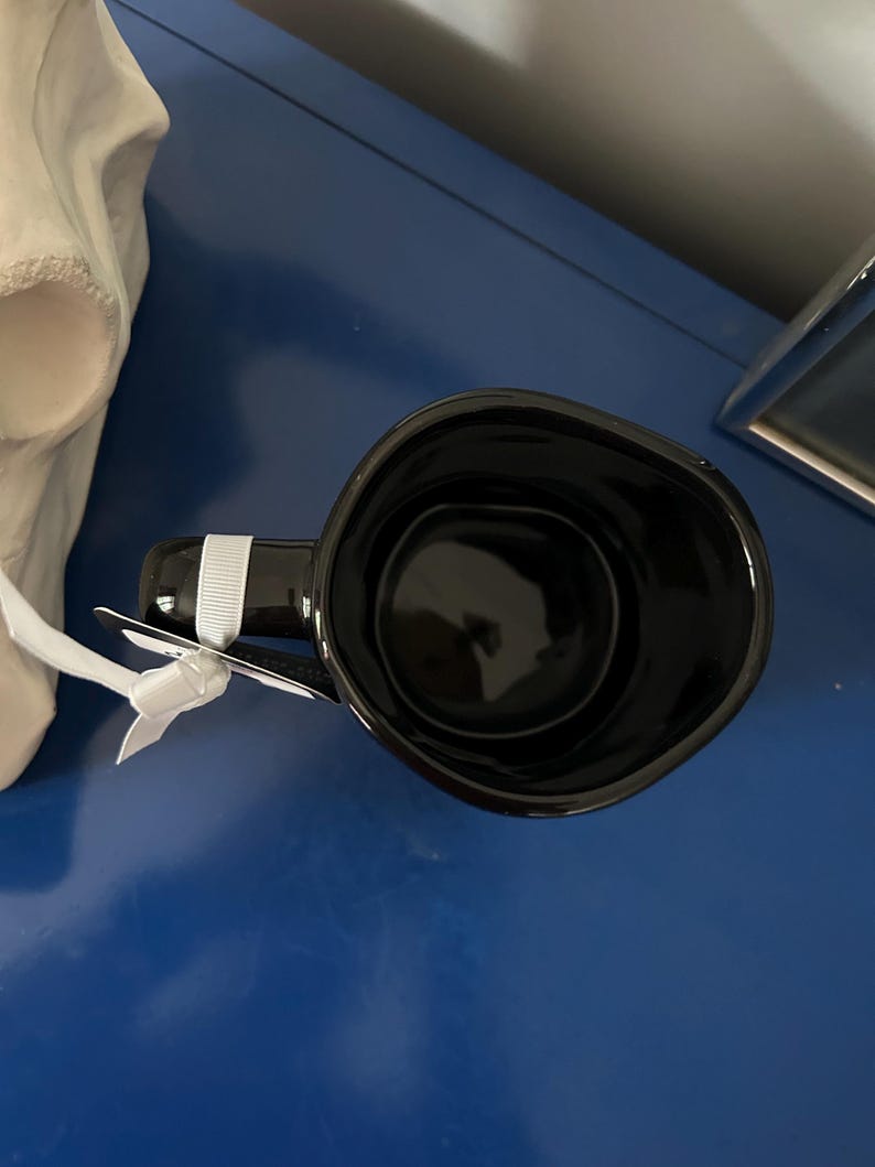 Op de afbeelding: Een zwarte keramische mok met een glanzende afwerking, direct van bovenaf bekeken. De mok heeft een enkel handvat met een wit lint eromheen gebonden en een donkere, lege binnenkant. De rand van de mok heeft een licht onregelmatige, ronde vorm.