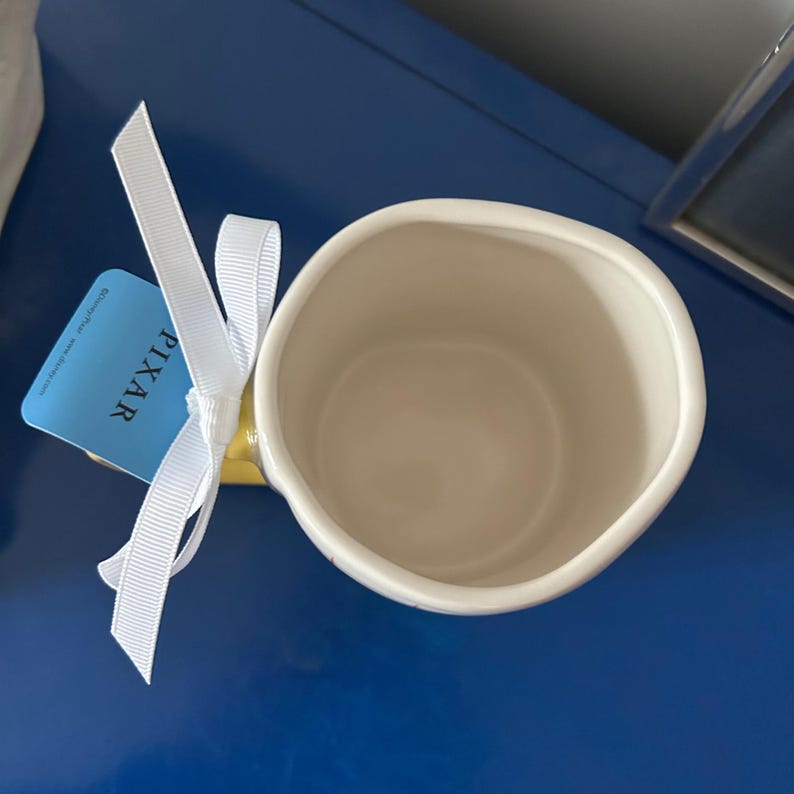 May include: A white ceramic mug with a yellow rim and a white ribbon tied around the handle. The mug is sitting on a blue surface. The tag on the ribbon says "PIXAR".