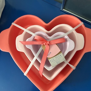 May include: A set of three heart-shaped bakers in shades of pink and coral, tied together with white plastic and a pink ribbon. A tag reads "RAE DUNN ARTISAN COLLECTION BY MAGENTA Set of 3 Bakers."