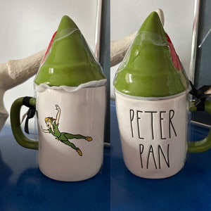 Pode incluir: Uma caneca de cerâmica branca com uma tampa verde em forma de chapéu do Peter Pan. A caneca apresenta uma imagem de desenho animado do Peter Pan voando e o texto "PETER PAN" na frente.