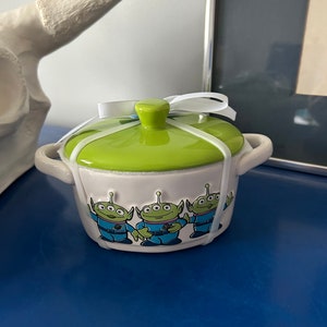 May include: A small, ceramic, lidded dish with a light green lid and a white ribbon tied around it. The dish is light pink with three cartoon alien characters in blue uniforms. The dish has two small handles.