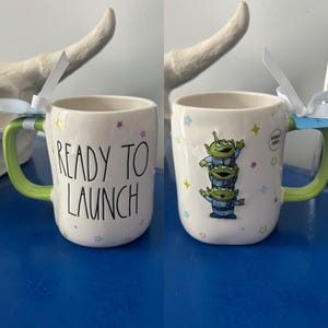 May include: White ceramic mug with a green handle, featuring the text "READY TO LAUNCH" on one side and a stack of three green alien characters on the other. The mug is decorated with colorful star accents.