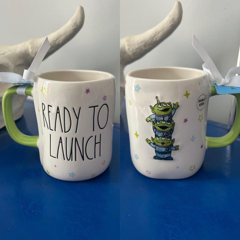 May include: White ceramic mug with a green handle, featuring the text "READY TO LAUNCH" on one side and a stack of three green alien characters on the other. The mug is decorated with colorful star accents.