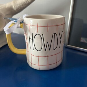 May include: White ceramic mug with a red and white checkered pattern and the word "HOWDY" in black lettering. The mug has a yellow handle.