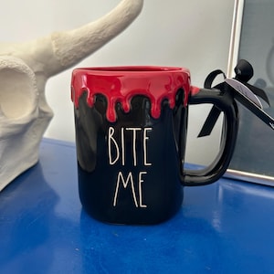 Puede incluir: Taza de cerámica negra con borde rojo goteando. La taza tiene un asa negra y las palabras "BITE ME" en letras blancas. Una cinta negra está atada al asa.
