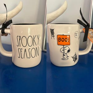 Könnte beinhalten: Eine weiße Keramiktasse mit schwarzem Innenraum und Griff. Die Tasse zeigt auf der einen Seite die Worte "SPOOKY SEASON" und auf der anderen Seite eine Snoopy-Grafik, die ein "BOO!"-Schild hält. Schwarze Spinnweben sind ebenfalls auf der Tasse. Ein schwarzes Band ist am Griff befestigt.