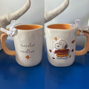 Könnte beinhalten: Ein weißer Keramikbecher mit orangefarbenem Innenraum und Henkel, an dem ein weißes Band befestigt ist. Eine Seite zeigt den Schriftzug "sweater weather" mit einem kleinen Blatt, die andere Seite ein erhabenes Snoopy-Motiv in einem Pullover.