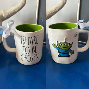 May include: White ceramic mug with a green rim and a black and white graphic that says "Prepare to be Chosen". The other side of the mug features a green alien character with three eyes, wearing a blue jumpsuit and a black belt. The alien is waving with its right hand.
