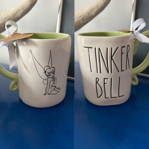 May include: White ceramic mug with a green interior and handle. The mug features a black and white illustration of Tinker Bell on one side and the words "TINKER BELL" on the other. A white ribbon is tied around the handle.