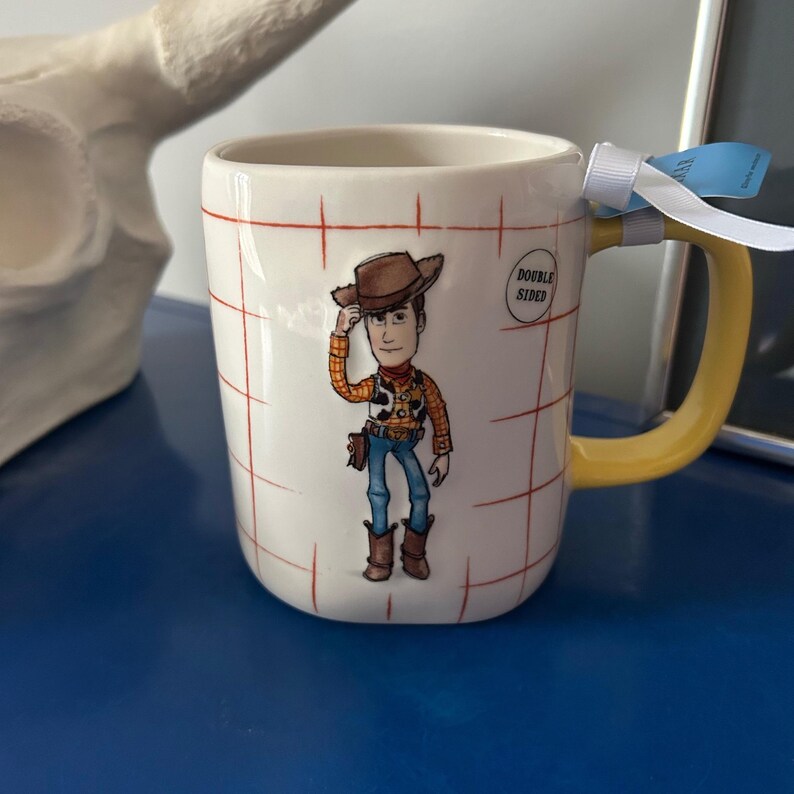 May include: White ceramic mug with a red and white grid pattern. The mug features a cartoon illustration of Woody from Toy Story, wearing his signature cowboy hat and vest. The mug has a yellow handle and a sticker that says "Double Sided".