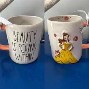 May include: White ceramic mug with a pink handle. The mug has a black graphic that says "Beauty is found within". The other side of the mug features a cartoon image of Belle from Beauty and the Beast.