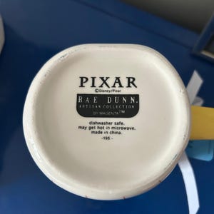 May include: White ceramic mug with a red and blue design. The bottom of the mug has the text "PIXAR", "Disney/Pixar", "RAE DUNN.", "ARTISAN COLLECTION", "BY MAGENTATM", "dishwasher safe. may get hot in microwave. made in china. -195-".