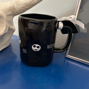 Op de afbeelding: Een zwarte keramische mok met een witte afbeelding van een schedelgezicht, de tekst EEK! en het woord NIGHTMARE. De mok heeft een gebogen handvat met een wit lint eromheen gestrikt en een zwart label bevestigd met een wit touwtje.