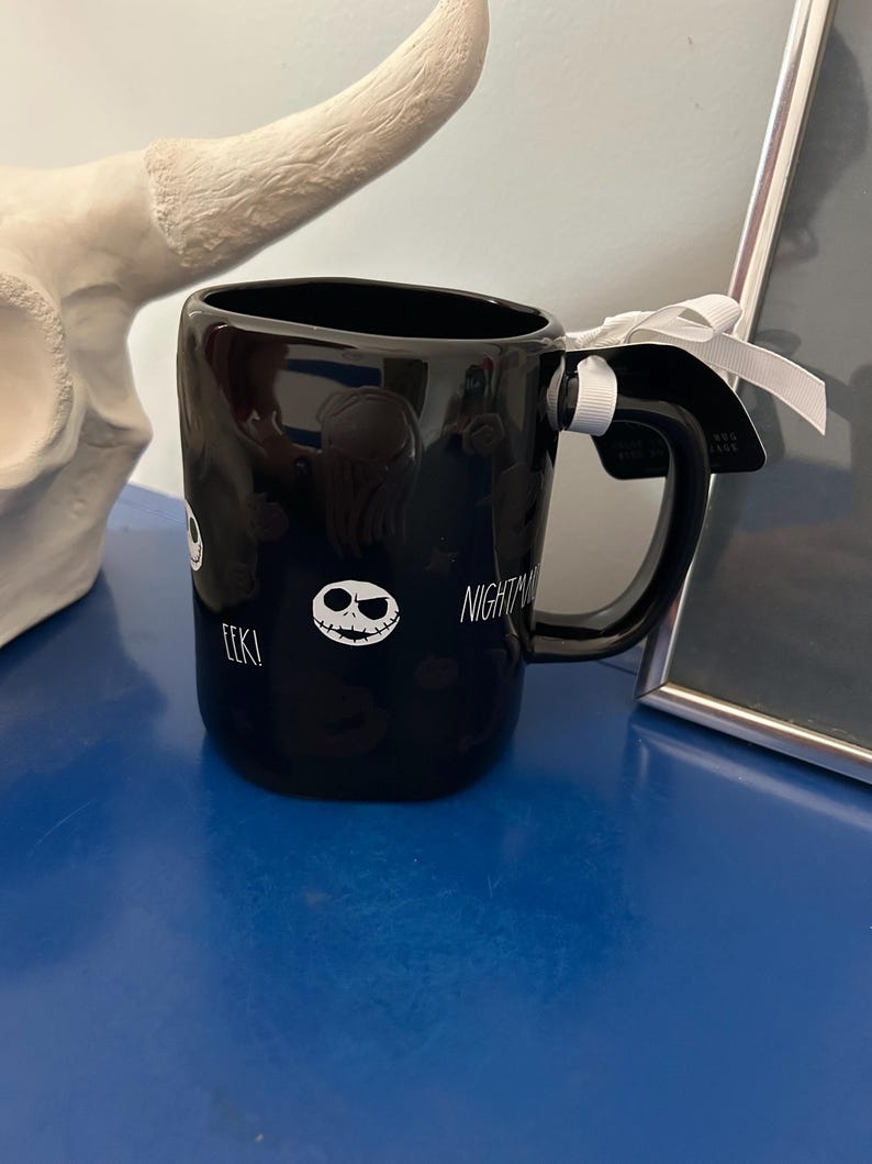 Op de afbeelding: Een zwarte keramische mok met een witte afbeelding van een schedelgezicht, de tekst EEK! en het woord NIGHTMARE. De mok heeft een gebogen handvat met een wit lint eromheen gestrikt en een zwart label bevestigd met een wit touwtje.