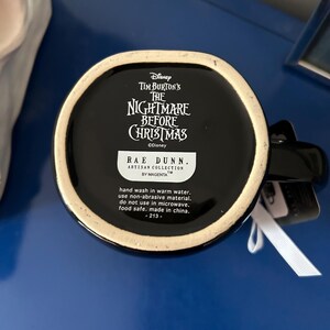 Op de afbeelding: Een zwarte keramische mok met het Disney Tim Burton's The Nightmare Before Christmas logo op de onderkant. Een wit label toont de tekst Rae Dunn Artisan Collection by Magenta, met onderhoudsinstructies en de herkomst made in China.