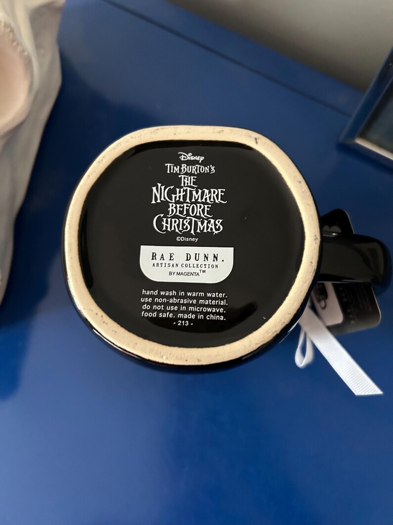 Op de afbeelding: Een zwarte keramische mok met het Disney Tim Burton's The Nightmare Before Christmas logo op de onderkant. Een wit label toont de tekst Rae Dunn Artisan Collection by Magenta, met onderhoudsinstructies en de herkomst made in China.