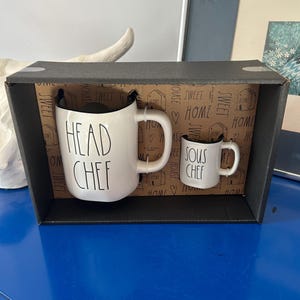 以下が含まれることがあります： 「HEAD CHEF」と「SOUS CHEF」の文字が黒で書かれた白いセラミックマグのセット。マグカップは、「SWEET HOME」の文字が入った装飾的な内装の黒い箱に展示されています。