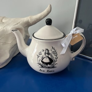 Puede incluir: Tetera de cerámica blanca con borde, asa y pomo de tapa negros. La tetera presenta una ilustración en blanco y negro de Alicia en el país de las maravillas con la frase "tea time". Una etiqueta de Disney está adjunta.